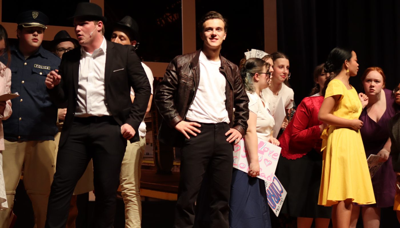 Conrad Birdie's actor posing with his hands on his hips surrounded by other characters